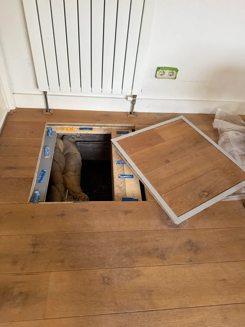 Open kruipluik in een houten vloer, met een medewerker van PuinVrij die de kruipruimte in gaat