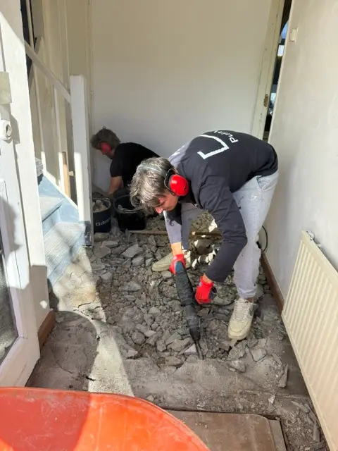 Twee medewerkers van PuinVrij breken de tegelvloer in een hal op, met puinbrokken en emmers op de vloer